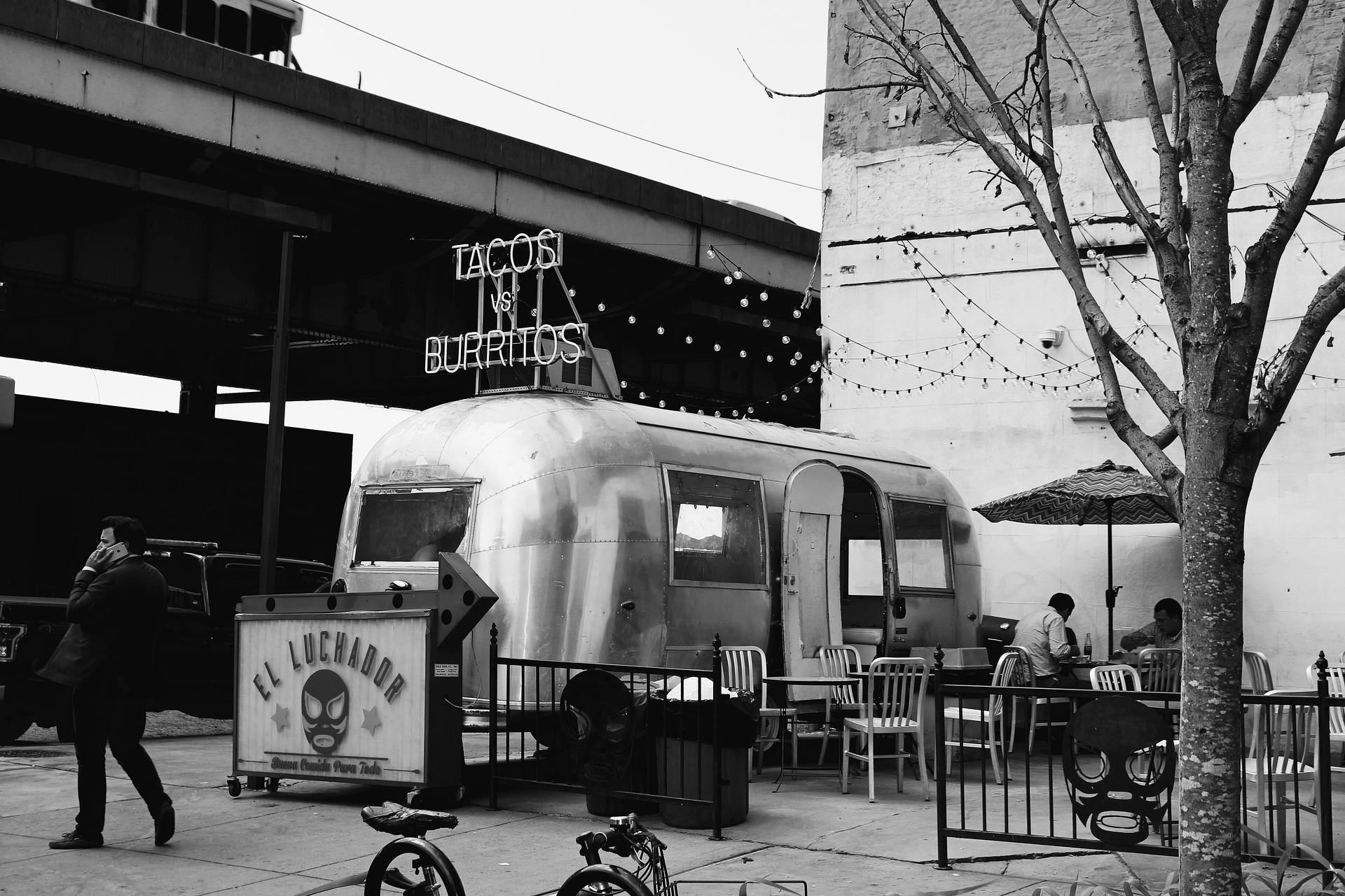 Das Schwarz-weiß Bild zeigt einen alten silbernen Foodtruck, welcher Tacos und Burritos verkauft. Um dem Truck herum befinden sich Stühle und Tische zum Essen.